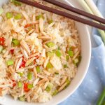 A bowl of pineapple rice topped with chopped green onions and, slivered almonds, with a pair of chopsticks resting on top. A blue napkin and green onions are in the background.