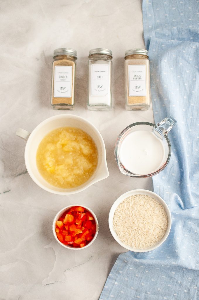 A flat lay photo shows jars of ginger, salt, and pepper, alongside bowls of crushed pineapple, coconut milk, uncooked rice, and diced red bell pepper on a marble surface with a blue cloth.