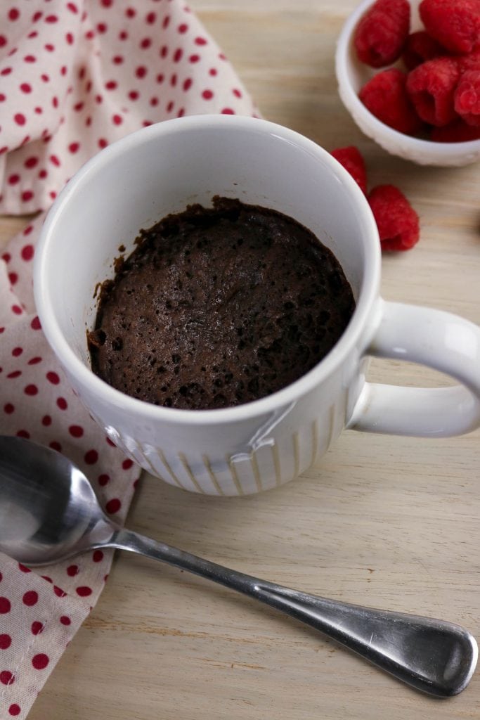 A chocolate mug cake sits in a white mug on a wooden table, next to a silver spoon, a red polka dot cloth, and a bowl of fresh raspberries.