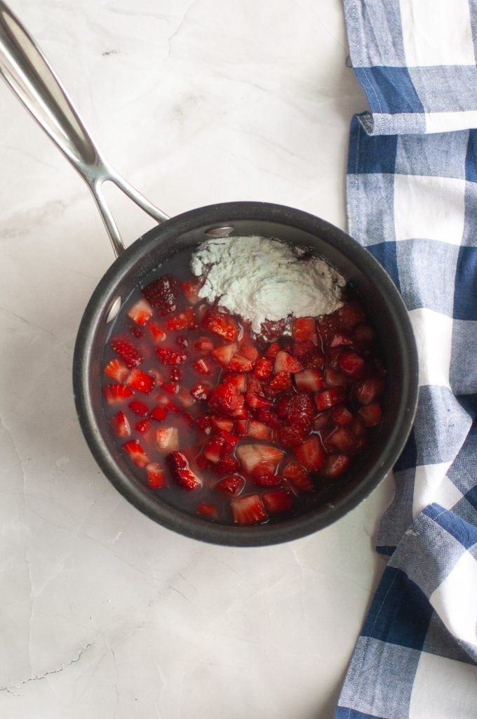 A saucepan filled with chopped strawberries, a white powder, and liquid sits on a white surface next to a blue and white checkered cloth.