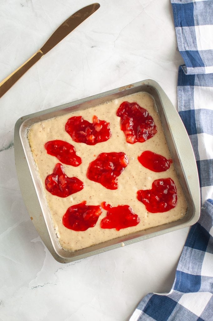 A square metal baking pan filled with cake batter topped with dollops of red jam sits on a marble surface next to a blue and white checkered cloth and a butter knife.