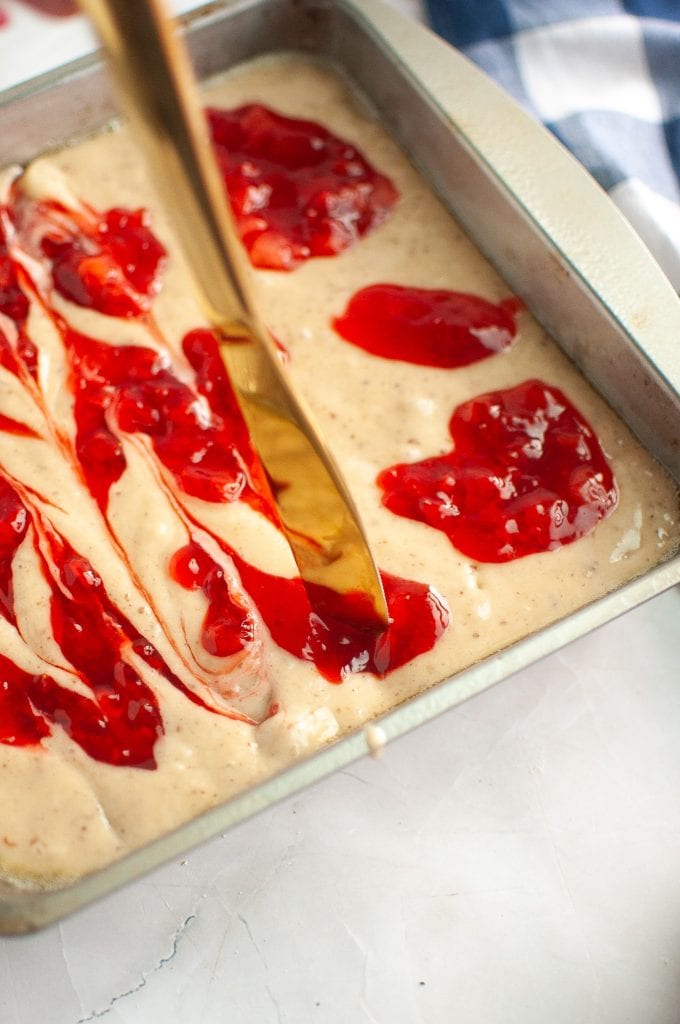A close-up of a metal baking pan filled with cake batter, topped with dollops of red fruit jam being swirled with a gold knife. A blue-and-white cloth is partially visible in the background.