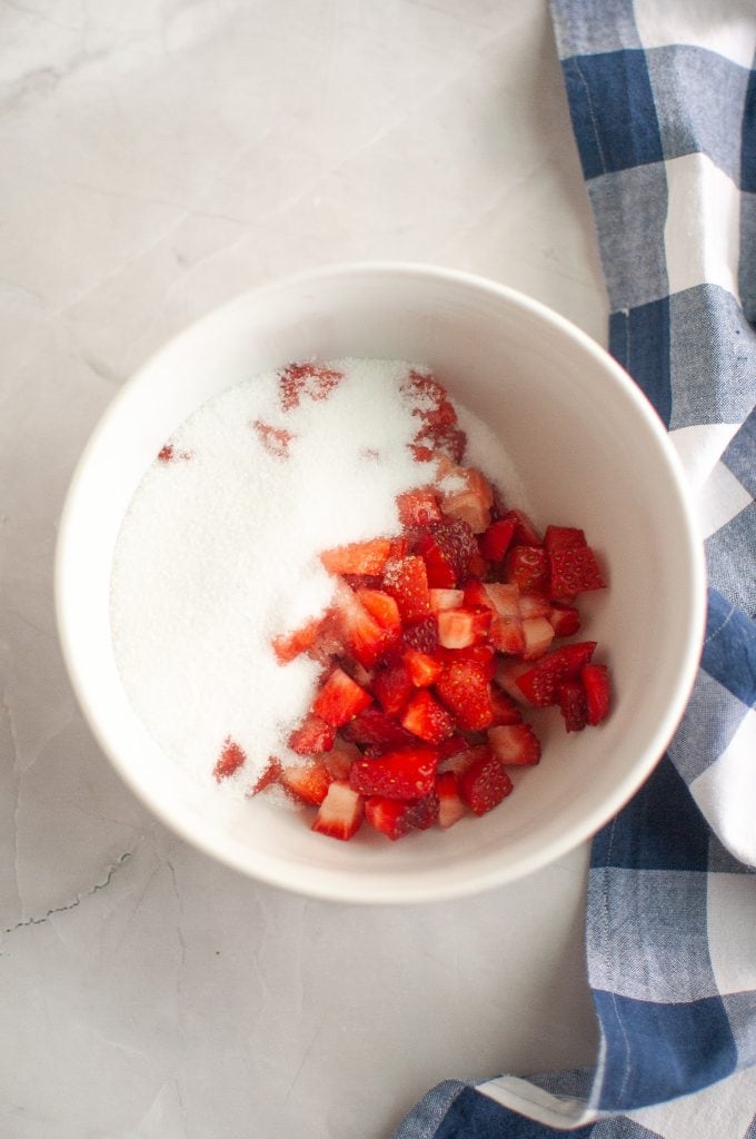 A white bowl filled with chopped strawberries and granulated sugar sits on a light countertop next to a blue and white checkered cloth.