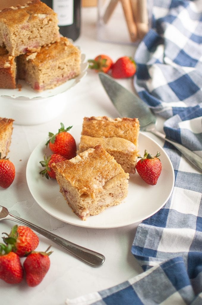 Two slices of vegan sourdough strawberry cake on a white plate with fresh strawberries, next to a stack of cake squares on a stand. A blue and white checkered cloth and utensils are arranged nearby.