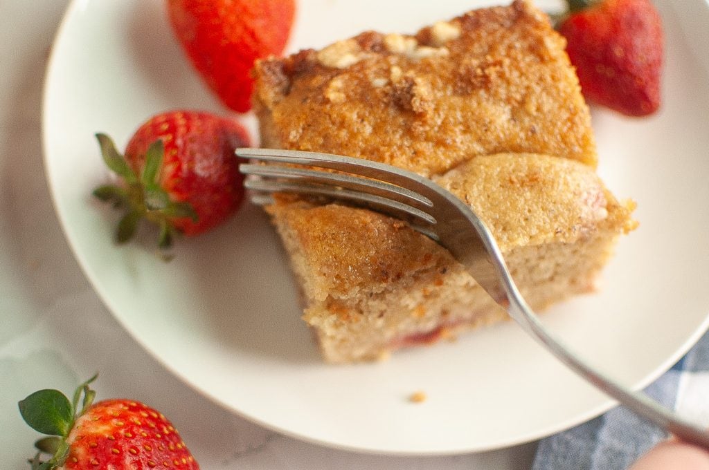 A fork cuts into a slice of vegan sourdough strawberry cake on a white plate, surrounded by fresh strawberries.