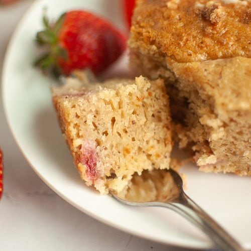 A close-up of a fork holding a piece of moist, crumbly vegan sourdough strawberry cake with visible strawberry pieces, on a white plate with whole strawberries in the background.