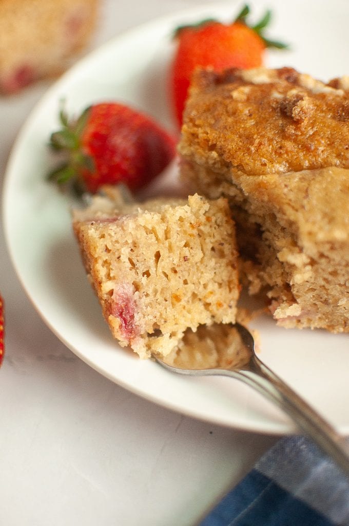 A close-up of a fork holding a piece of moist, crumbly vegan sourdough strawberry cake with visible strawberry pieces, on a white plate with whole strawberries in the background.