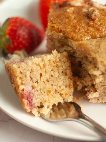 A close-up of a plate with moist, golden vegan sourdough strawberry cake, a piece broken off with a fork, and fresh strawberries surrounding the cake on a white surface.