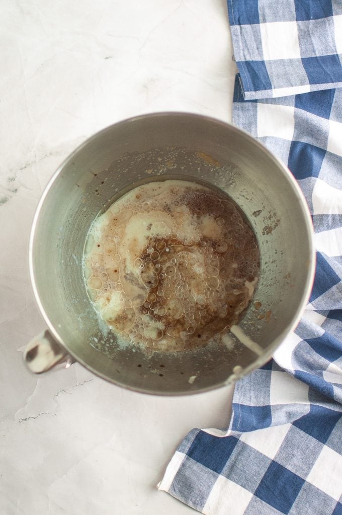 A metal mixing bowl with wet and dry baking ingredients, including bubbles and foam on top, sits on a white surface next to a blue and white checkered kitchen towel.